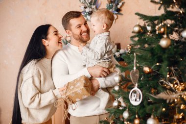 Genç bir aile Noel 'i birlikte kutluyor. Esmer anne, sakallı baba ve sarışın oğul Noel ağacının yanında duruyor ve bir hediye kutusu tutuyorlar. Bej örgülü giysiler giyen bir aile..