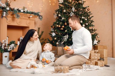 Genç bir aile Noel 'i birlikte kutluyor. Esmer anne, sakallı baba ve sarışın oğul Noel ağacının yanında oturup mandalina tutuyorlar. Bej örgülü giysiler giyen bir aile..