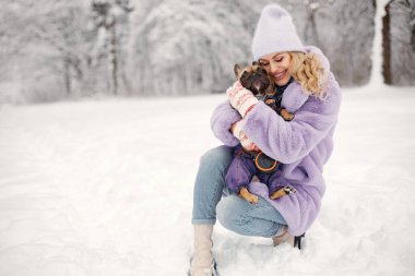 Genç sarışın kadın ve köpeği buldok kışın karda oynuyorlar. Mor, kalın ceket ve örgü şapka giyen beyaz bir kadın. Sıcak tulum giymiş bir köpek..