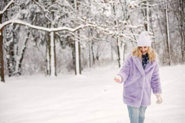Genç sarışın bir kadının portresi, kışın karın üzerinde duruyor. Beyaz kadın mor, kalın kürk manto ve örgü şapka giyiyor. Kadın fotoğraf için poz veriyor..