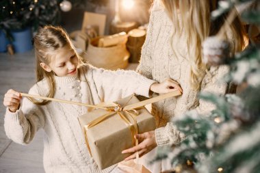 Evde Noel ağacının yanında oturan küçük bir kızın kesilmiş fotoğrafı. Elinde hediye kutusu tutan bir kız. Beyaz ve bej elbiseli kadın ve kız.