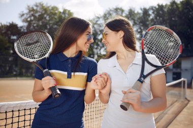 Tenis kortunda raket tutan iki kız. Beyaz ve mavi spor elbiseler ve güneş gözlüğü takan kadınlar. Kızlar bir arada durup poz veriyorlar..