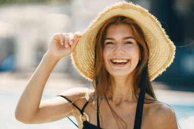 Yüzme havuzunun yanında duran yaz şapkalı genç kız. Siyah mayo ve şapka giyen bir kız. Havuz kenarında poz veren bir kız..