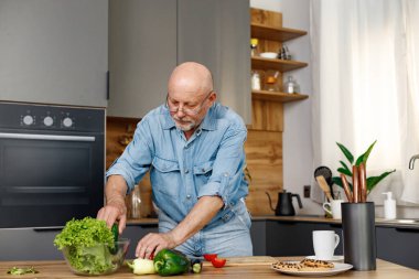 Yeşil salata, biber ve domateslerden yemek pişiren emekli son sınıf öğrencisi. Adam mutfağın içinde duruyor. Kot pantolon ve gömlek giyen adam..
