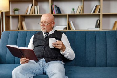 Kıdemli adam kanepede oturuyor ve akşamları evde kitap okuyor. Çay içen bir adam. Beyaz gömlek ve siyah yelek giyen adam..