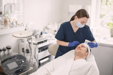 Adam kozmetolog kabinesinde yatıyor ve yüz derisi için bir prosedürü var. Mavi tıbbi kostüm ve yüz maskesi takan kadın kozmetik uzmanı. Beyaz bornoz giyen adam..