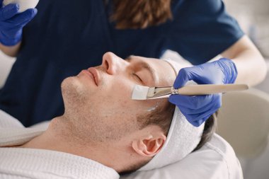 Adam kozmetolog kabinesinde yatıyor ve yüz derisi için bir prosedürü var. Mavi tıbbi kostüm giyen tanınmamış bir kadın kozmetik uzmanı. Erkek yüzüne maske takan bir kadın..