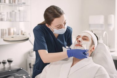 Adam kozmetolog kabinesinde yatıyor ve yüz derisi için bir prosedürü var. Mavi tıbbi kostüm ve yüz maskesi takan kadın kozmetik uzmanı. Erkek yüzüne maske takan bir kadın..