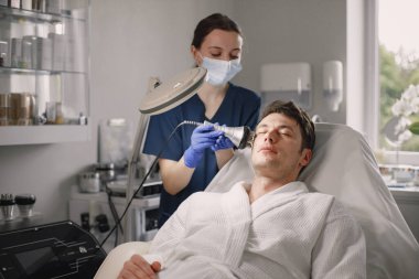 Kozmetolog kabinesinde yatan genç bir adam ve yüz derisi için bir çürüme prosedürü var. Mavi tıbbi kostüm ve yüz maskesi takan kadın kozmetik uzmanı. Beyaz bornoz giyen adam..