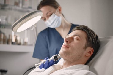 Kozmetolog kabinesinde yatan genç bir adam ve yüz derisi için bir çürüme prosedürü var. Mavi tıbbi kostüm ve yüz maskesi takan kadın kozmetik uzmanı. Beyaz bornoz giyen adam..