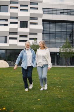 Yazın çimlerin üzerinde duran Kafkasyalı yaşlı çift. Erkek ve kadın yürüyor ve el ele tutuşuyorlar. Beyaz tişörtlü kadın ve mavi erkek..