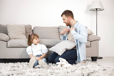 Sevgi dolu baba, Down sendromlu kızıyla evde birlikte oynuyor. Kanepenin yanında bir adam ve kız oturuyor. Sakallı adam mavi gömlekli ve kız beyaz gömlekli..