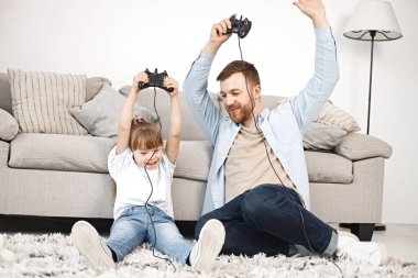 Sevgi dolu baba, Down sendromlu kızıyla evde birlikte oynuyor. Kız ve erkek kanepede oturup joystickleri tutuyorlar. Sakallı adam mavi gömlekli ve kız beyaz gömlekli..