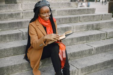 Uzun saç stili olan genç siyahi bir kadın elinde kitapla merdivenlerde oturuyor. Kahverengi ceketli, turuncu eşarplı ve siyah şapkalı bir kadın. Gözlüklü kadın sonbaharda dışarıda kitap okuyor..