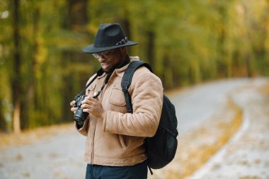 Genç siyahi bir adam ormanda elinde kamerayla dikiliyor. Ormanda yürüyen erkek fotoğrafçı. Kahverengi ceketli, siyah şapkalı ve sırt çantalı bir adam..