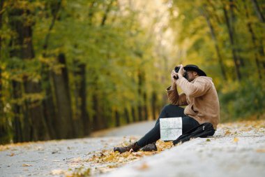 Ormanda kameralı genç bir zenci yolda oturuyor. Ormanda bir erkek fotoğrafçı. Kahverengi ceketli, siyah şapkalı ve sırt çantalı bir adam..