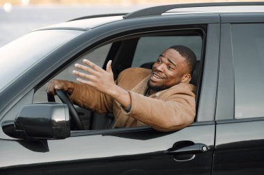 Arabasında oturan sinirli genç siyahi adamın yakın plan portresi. Sakallı, kahverengi ceketli bir adam. Erkek sürücü gergin görünüyor..