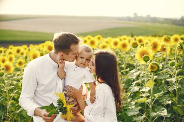 Güneşli bir günde, ayçiçeği tarlasında genç bir ailenin fotoğrafı. Esmer anne, baba ve küçük sarışın oğulları fotoğraf için poz veriyorlar. Oğlu elinde tutan adam.