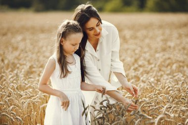 Güneşli bir günde, buğday tarlasında beyaz elbiseli genç bir anne ve kızının fotoğrafı. Esmer anne ve küçük kızı fotoğraf için poz veriyorlar. Buğday sapına bakan kız.