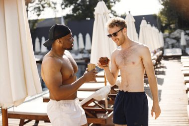 Beyaz ve siyah Amerikalı çocuklar yüzme havuzunun yanında dinleniyorlar. Beyaz ve siyah mayo ve güneş gözlüğü takan çocuklar. Kokteyl içen adamlar.