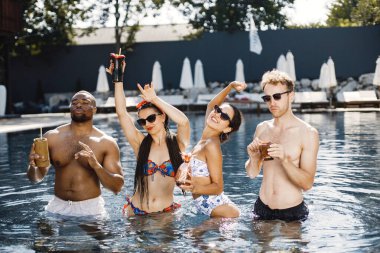 İki genç kız ve iki erkek arkadaşları yüzme havuzunda dinleniyorlar. Mavi ve kırmızı mayo giyen kızlar. Siyah Amerikalı erkek, beyaz erkek ve kızlar havuzda durup kokteyl içiyorlar..