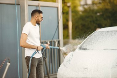 Moda elbiseli genç adam araba yıkama istasyonunda yüksek basınçlı sabun makinesiyle araba yıkıyor. Beyaz tişört ve kot pantolon giyen bir adam. Hintli adam beyaz arabasını yıkıyor..