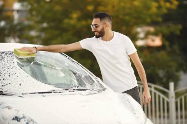 Moda elbiseli genç adam araba yıkama istasyonunda çamaşır bezi kullanarak araba yıkıyor. Beyaz tişört ve kot pantolon giyen bir adam. Hintli adam beyaz arabasını yıkıyor..