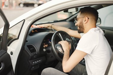 Gözlüklü genç adam arabasının içinde halıyla parlıyor. Beyaz tişörtlü bir adam. Hintli adam arabasını siliyor..