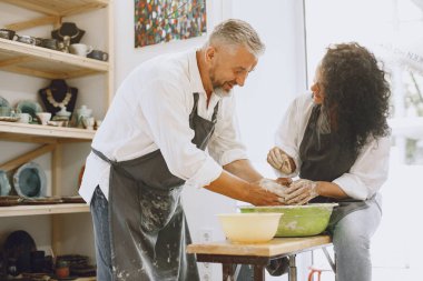 Ortak yaratıcı çalışma. Günlük giysiler ve önlükler içinde yetişkin, zarif bir çift. İnsanlar kilden bir stüdyoda çömlek çarkında kase yapıyorlar..