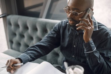 Kahve dükkanında oturup latte içen siyahi bir adam. Güneş gözlüklü ve gri gömlekli bir adam. İş adamı telefon kullanıyor..