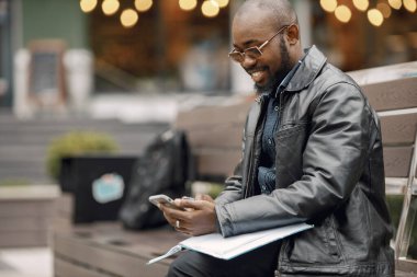 Şehirdeki bir bankta oturan siyah bir adam. Güneş gözlüklü ve deri ceketli bir adam. İş adamının evrakları var ve telefon kullanıyor..