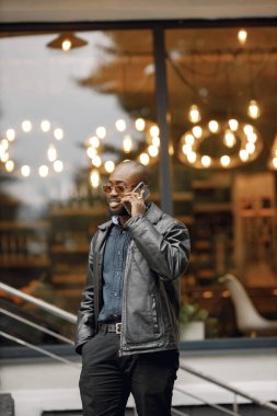 Şehirdeki bir caddede duran siyah adam. Güneş gözlüklü ve deri ceketli bir adam. İş adamı telefon kullanıyor..