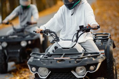 Focus on a woman riding an ATV quad bikes in autumn forest. Two friends maneuvering off-road ATV. Couple wearing grey sportive costumes.