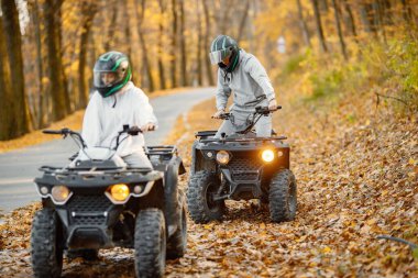 A young caucasian man and woman riding an ATV quad bikes in autumn forest. Two friends maneuvering off-road ATV. Couple wearing grey sportive costumes.
