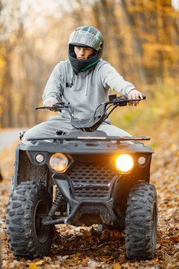 A young caucasian man in helmet riding an ATV quad bike in autumn forest. Boy maneuvering off-road ATV. Man wearing grey sportive costume.