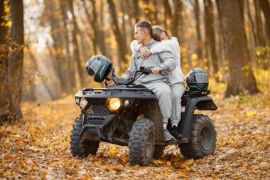 A young caucasian man and woman riding an ATV quad bike in autumn forest. Lovely couple hugging. Couple wearing grey sportive costumes.