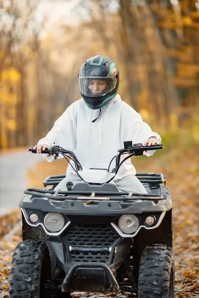 A young caucasian woman in helmet riding an ATV quad bike in autumn forest. Girl maneuvering off-road ATV. Woman wearing grey sportive costume.