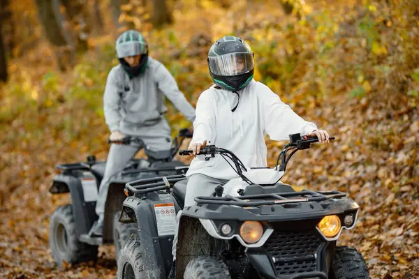 A young caucasian man and woman riding an ATV quad bikes in autumn forest. Two friends maneuvering off-road ATV. Couple wearing grey sportive costumes.