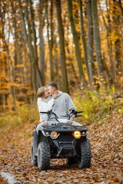 A young caucasian man and woman riding an ATV quad bike in autumn forest. Lovely couple kissing. Couple wearing grey sportive costumes.