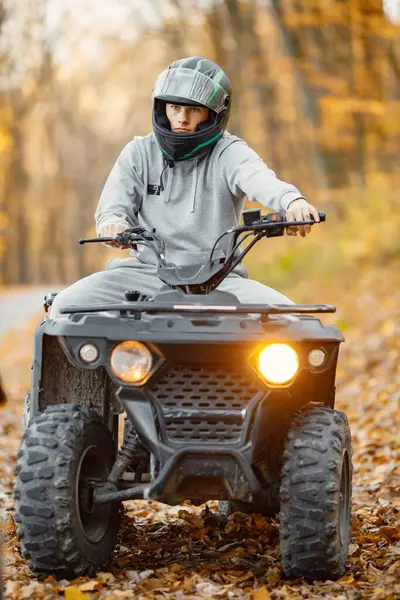 A young caucasian man in helmet riding an ATV quad bike in autumn forest. Boy maneuvering off-road ATV. Man wearing grey sportive costume.