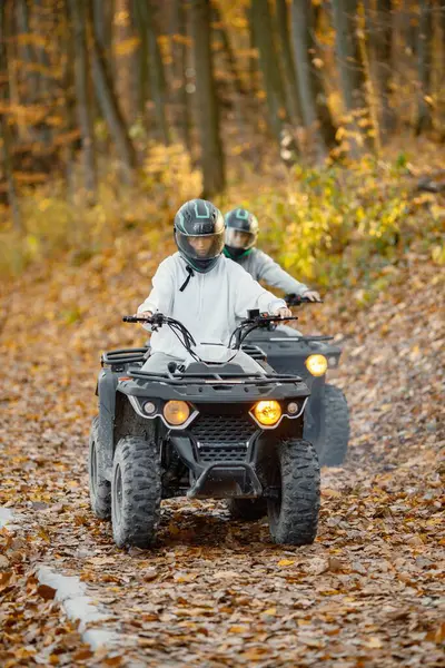 A young caucasian man and woman riding an ATV quad bike in autumn forest. Couple maneuvering off-road ATV. Couple wearing grey sportive costumes.