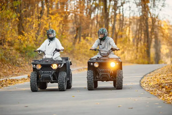 A young caucasian man and woman riding an ATV quad bikes in autumn forest. Couple maneuvering off-road ATV. Couple wearing grey sportive costumes.