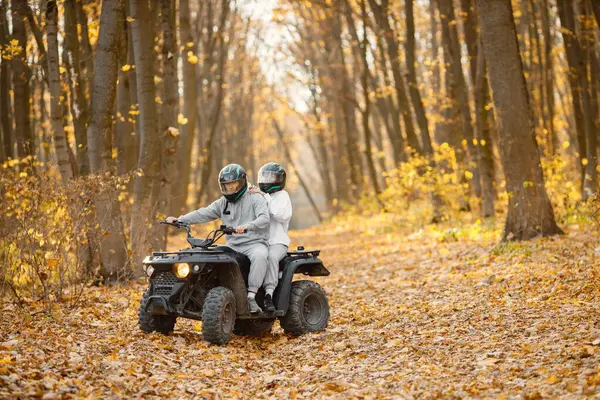 A young caucasian man and woman riding an ATV quad bike in autumn forest. Couple maneuvering off-road ATV. Couple wearing grey sportive costumes.