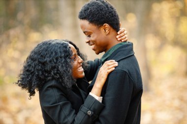 Siyah genç adam ve kız arkadaşı sarılıyor. Sonbahar parkında yürüyen romantik bir çift. Siyah paltolu kadın ve erkek..