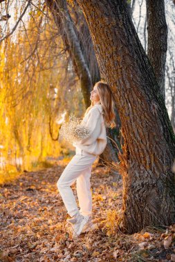 Young blonde girl with flowers standing in autumn park near the lake. Woman wearing beige sweater and white trousers. Girl posing for a photo in sunny day.