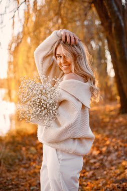 Young blonde girl with flowers standing in autumn park near the lake. Woman wearing beige sweater. Girl posing for a photo in sunny day.