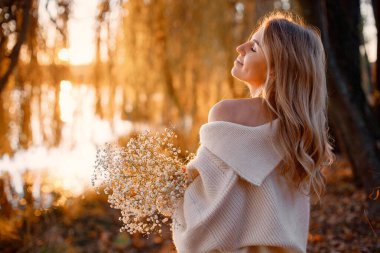 Young blonde girl with flowers standing in autumn park near the lake. Woman wearing beige sweater. Girl posing for a photo in sunny day.
