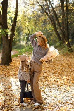 Anne ve küçük kızı sonbahar yürüyüşünde oynayıp gülüyorlar. Annem el işi kağıdında ekmek tutuyor. Gri kazak giyen kadın ve kız.