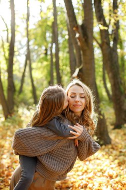Anne ve küçük kızı sonbahar yürüyüşünde birbirlerine sarılıyorlar. Kızını sırtında taşıyan bir kadın. Gri kazak giyen kadın ve kız.