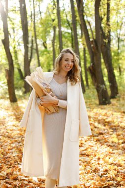 Kıvırcık genç bir kadının sonbaharda el sanatları kağıdında baget tutarken portresi. Parkta sıcak sonbaharın tadını çıkaran bir kadın. Bej elbiseli ve beyaz ceketli kadın..